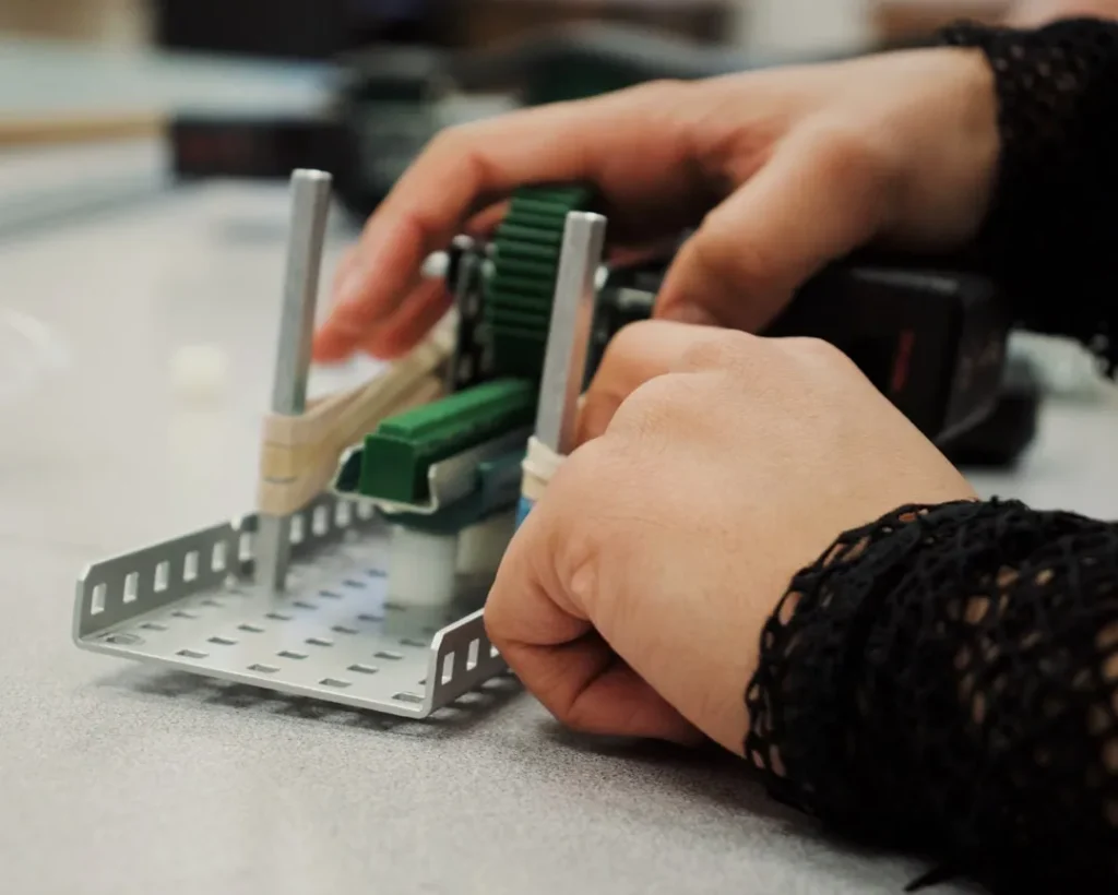 Hands assembling a combat robot component with green parts and metal frame in a robotics workshop setting.