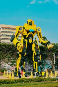 Large yellow combat robot statue in outdoor setting, resembling Project Titan, surrounded by caution cones and urban backdrop, emphasizing engineering and robotics themes.